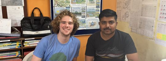 AIF Banyan Impact Fellow Mukund Michael Adamson (left) and CEE Jasdan Engineer Ashok Sapara (right) posing for a photo at Ashok Bhai’s desk in the CEE Jasdan field office. Scientific diagrams dot the background along with intervention files.