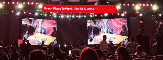 Audience seated in a large conference hall watching a stage with multiple big screens playing an adevertising video of a company's inclusive work video; a red banner reads “Great Place To Work For All Summit Mumbai 2026.”